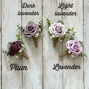 Op de afbeelding: Vier florale corsages met schuimrozen in de kleuren pruim, donkerlavendel, lichtlavendel en lavendel. Elke corsage bevat groene eucalyptusbladeren, wit gipskruid en een steel omwikkeld met bruin touw, voorzien van tekstlabels voor elke kleur.