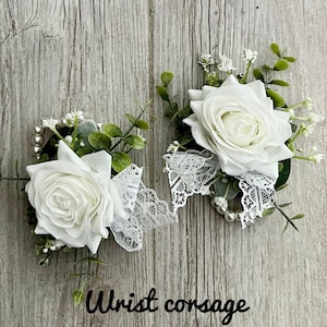 May include: Two white rose wrist corsages with lace and pearl accents. The corsages are on a wood background.