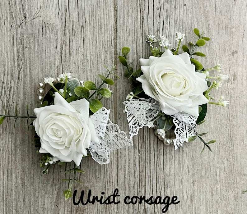 May include: Two white rose wrist corsages with lace and pearl accents. The corsages are on a wood background.