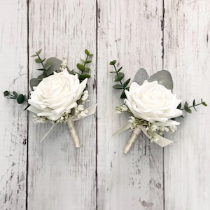 May include: Two white rose boutonnieres with greenery and white ribbon.