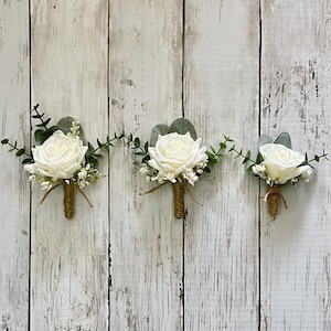 May include: Three white rose boutonnieres with greenery and twine-wrapped stems. The floral arrangements include small white flowers and green leaves. The boutonnieres are set against a white wooden background.