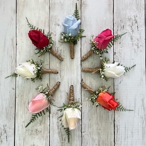 May include: Eight artificial rose buttonholes in various colours, including white, pink, red, coral, and blue. Each buttonhole is tied with twine and features small green leaves.