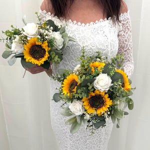 Peut inclure: Deux bouquets composés de tournesols, de roses blanches et de verdure. Les tournesols ont des pétales jaune vif et des centres brun foncé. Les bouquets sont enveloppés de ficelle. La personne porte une robe en dentelle blanche.