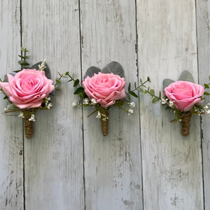 May include: Three pink rose boutonnieres with greenery and white baby's breath. Each boutonniere is tied with twine.