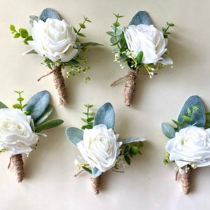 K&ouml;nnte beinhalten: F&uuml;nf Ansteckblumen mit wei&szlig;en Rosen, gr&uuml;nen Eukalyptusbl&auml;ttern und kleinen wei&szlig;en F&uuml;llblumen. Jeder Stiel ist mit braunem Bindfaden umwickelt. Die Blumenarrangements sind in einer Gruppe angeordnet und zeigen die wei&szlig;en Bl&uuml;tenbl&auml;tter.