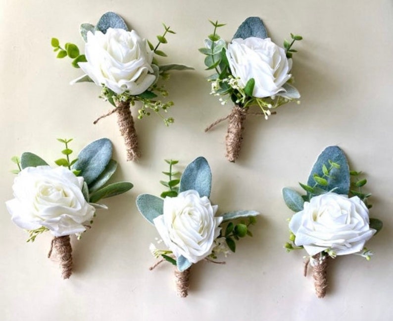 K&ouml;nnte beinhalten: F&uuml;nf Ansteckblumen mit wei&szlig;en Rosen, gr&uuml;nen Eukalyptusbl&auml;ttern und kleinen wei&szlig;en F&uuml;llblumen. Jeder Stiel ist mit braunem Bindfaden umwickelt. Die Blumenarrangements sind in einer Gruppe angeordnet und zeigen die wei&szlig;en Bl&uuml;tenbl&auml;tter.