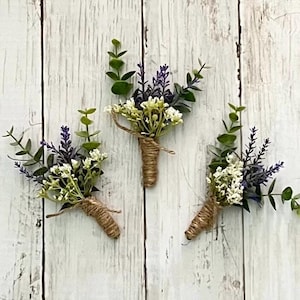 Op de afbeelding: Drie florale corsages met takjes paarse lavendel, kleine witte bloemetjes en groene eucalyptusbladeren. Elk arrangement is aan de basis samengebonden met natuurlijk bruin touw, wat zorgt voor een rustieke en samenhangende uitstraling.