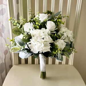 K&ouml;nnte beinhalten: Ein Blumenstrau&szlig; mit wei&szlig;en Rosen und wei&szlig;en Hortensienbl&uuml;ten, akzentuiert durch gr&uuml;ne Eukalyptusbl&auml;tter und weiches, salbeifarbenes Blattwerk. Die Stiele sind an der Basis fest mit einem glatten, wei&szlig;en Band umwickelt.