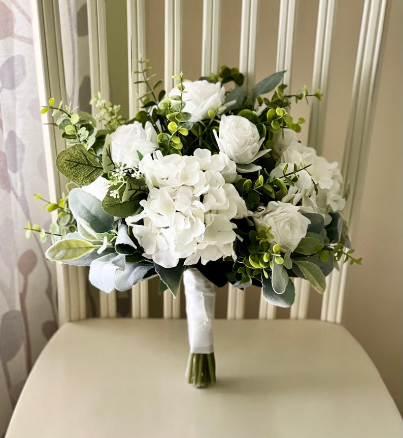 K&ouml;nnte beinhalten: Ein Blumenstrau&szlig; mit wei&szlig;en Rosen und wei&szlig;en Hortensienbl&uuml;ten, akzentuiert durch gr&uuml;ne Eukalyptusbl&auml;tter und weiches, salbeifarbenes Blattwerk. Die Stiele sind an der Basis fest mit einem glatten, wei&szlig;en Band umwickelt.