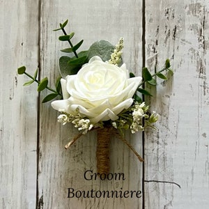 May include: A boutonniere featuring a single white rose, green eucalyptus leaves, and small white filler flowers. The stem is wrapped in brown twine, and the text Groom Boutonniere is printed in black at the bottom of the floral arrangement.