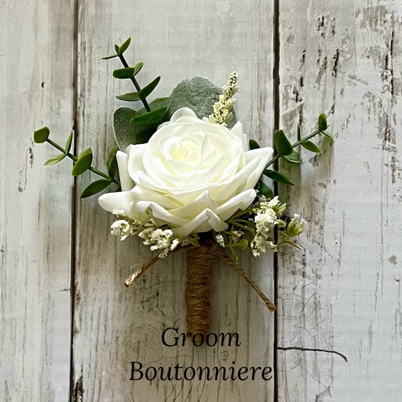 May include: A boutonniere featuring a single white rose, green eucalyptus leaves, and small white filler flowers. The stem is wrapped in brown twine, and the text Groom Boutonniere is printed in black at the bottom of the floral arrangement.