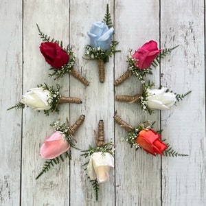 May include: Eight artificial rose buttonholes in various colours, including white, pink, red, coral, and blue. Each buttonhole is tied with twine and features small white flowers and greenery.