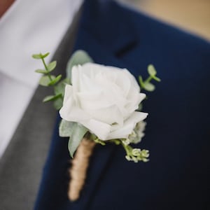 May include: A white rose boutonniere with green foliage and a brown twine wrap.