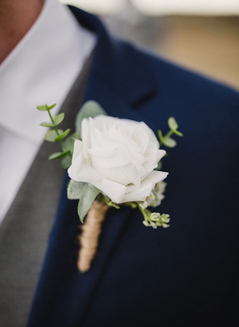 May include: A white rose boutonniere with green foliage and a brown twine wrap.