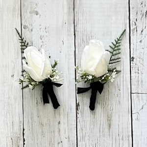 May include: Two white rose buttonholes with black ribbon accents and greenery.