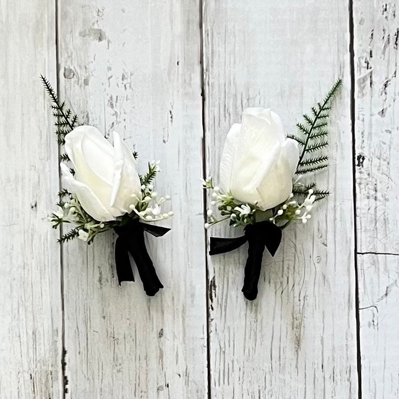May include: Two white rose buttonholes with black ribbon accents and greenery.