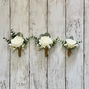 May include: Three boutonnieres featuring white roses, green eucalyptus leaves, and small white filler flowers. Each floral arrangement is accented with a brown twine-wrapped stem, creating a cohesive set of decorative floral accessories.