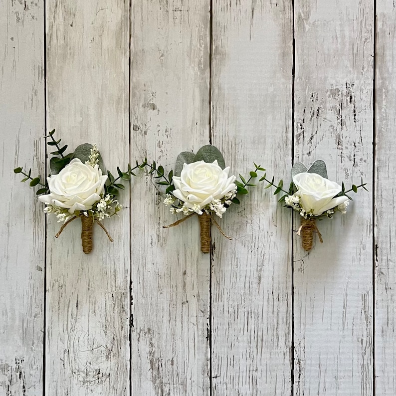 May include: Three boutonnieres featuring white roses, green eucalyptus leaves, and small white filler flowers. Each floral arrangement is accented with a brown twine-wrapped stem, creating a cohesive set of decorative floral accessories.