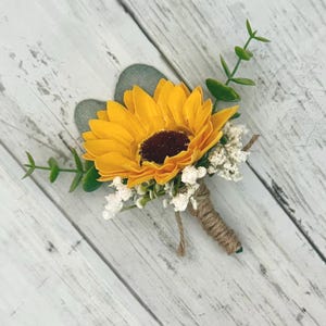 May include: A handcrafted boutonniere featuring a bright yellow sunflower with a dark brown center. The flower is accented with green leaves, small white flowers, and wrapped with twine. The boutonniere is set against a white wooden background.