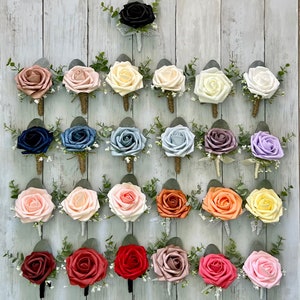 Op de afbeelding: Een collectie van vijfentwintig kunstmatige rozen corsages gerangschikt in vijf rijen. Elke corsage bevat een enkele schuimroos in diverse kleuren, geaccentueerd met groene eucalyptusbladeren, kleine witte vulbloemen en een omwikkelde steel.