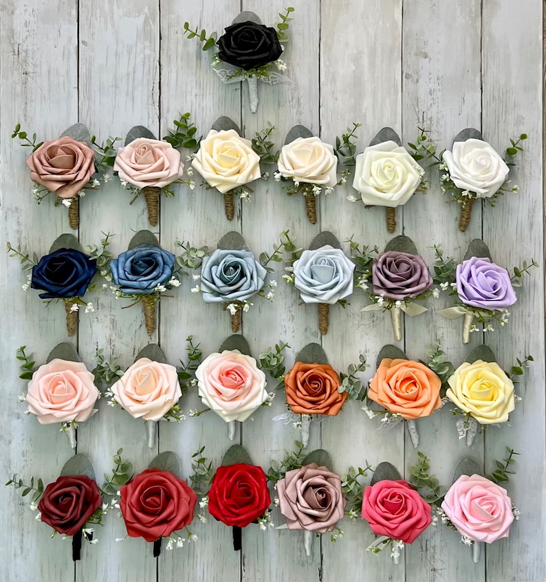 Op de afbeelding: Een collectie van vijfentwintig kunstmatige rozen corsages gerangschikt in vijf rijen. Elke corsage bevat een enkele schuimroos in diverse kleuren, geaccentueerd met groene eucalyptusbladeren, kleine witte vulbloemen en een omwikkelde steel.
