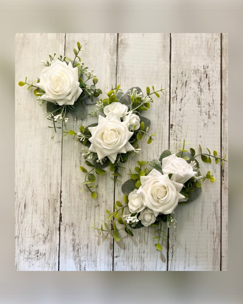 K&ouml;nnte beinhalten: Drei florale Ansteckblumen mit wei&szlig;en Rosen, kleinen wei&szlig;en F&uuml;llblumen und gr&uuml;nen Eukalyptusbl&auml;ttern. Jedes Arrangement besteht aus einer zentralen wei&szlig;en Rose, die von Gr&uuml;npflanzen und kleineren wei&szlig;en Bl&uuml;ten umgeben ist.
