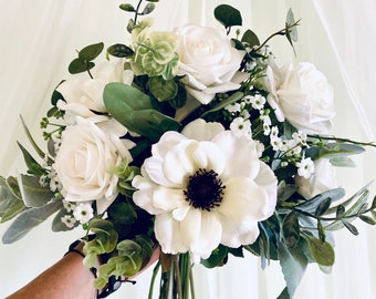 May include: A white bridal bouquet with white roses, white anemones, and greenery.