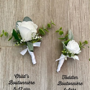 May include: Two white rose boutonni&egrave;res with greenery and white ribbon. The larger boutonni&egrave;re is labeled "Child's Boutonni&egrave;re 5-12 yrs" and the smaller boutonni&egrave;re is labeled "Toddler Boutonni&egrave;re 1-4yrs".