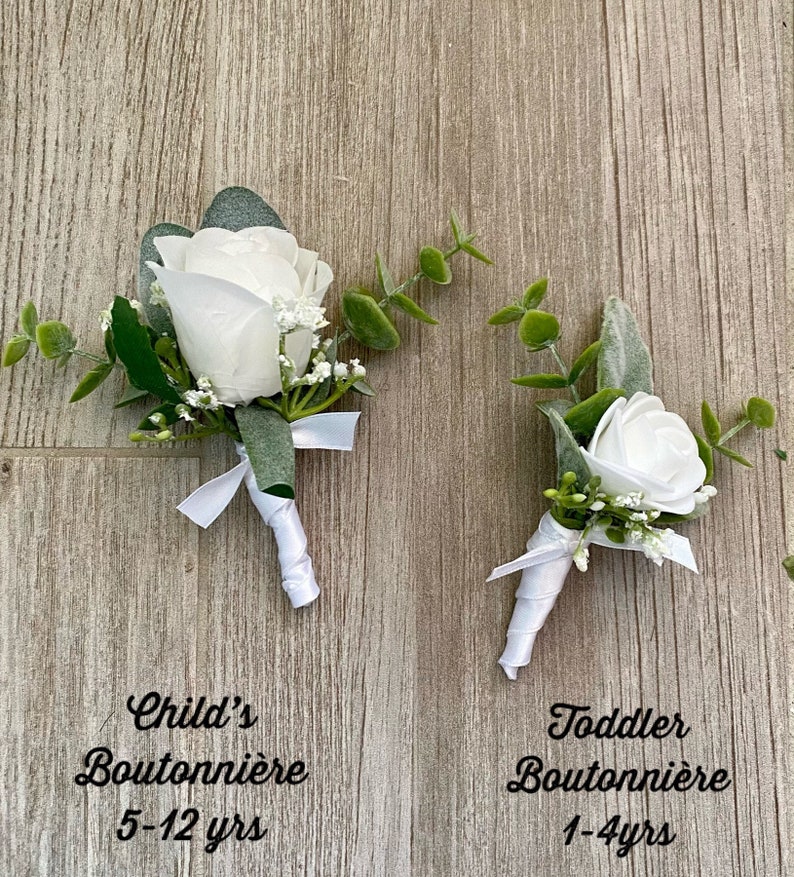 May include: Two white rose boutonni&egrave;res with greenery and white ribbon. The larger boutonni&egrave;re is labeled "Child's Boutonni&egrave;re 5-12 yrs" and the smaller boutonni&egrave;re is labeled "Toddler Boutonni&egrave;re 1-4yrs".