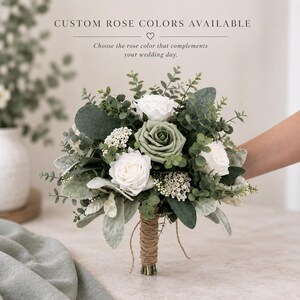 Op de afbeelding: Een bloemenboeket met witte en saliegroene rozen, kleine witte vulbloemen en diverse groene eucalyptusbladeren. De stelen zijn omwikkeld met natuurlijk bruin touw. De tekst bovenaan luidt: CUSTOM ROSE COLORS AVAILABLE.