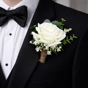 May include: A boutonniere featuring a single cream-colored rose, small white filler flowers, and green eucalyptus leaves. The stems are wrapped in brown twine, and the floral arrangement is pinned to the lapel of a black suit jacket with a white shirt.