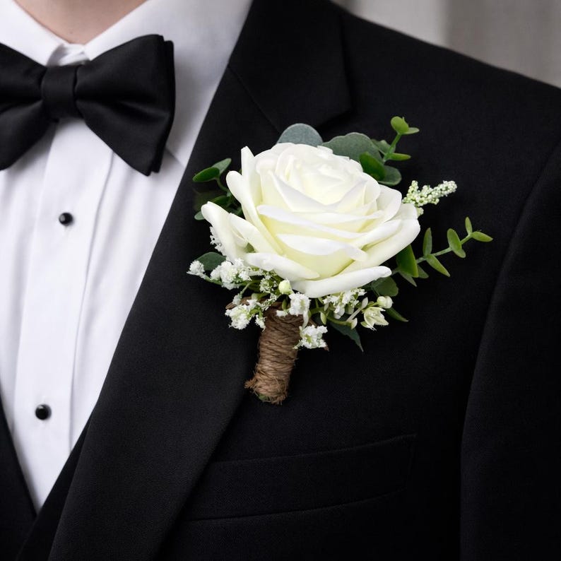 May include: A boutonniere featuring a single cream-colored rose, small white filler flowers, and green eucalyptus leaves. The stems are wrapped in brown twine, and the floral arrangement is pinned to the lapel of a black suit jacket with a white shirt.
