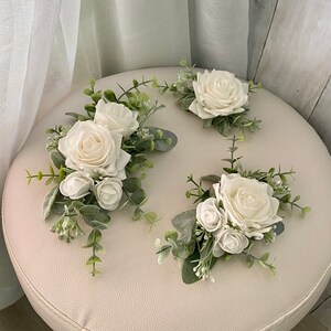 K&ouml;nnte beinhalten: Drei Blumenarrangements bestehend aus wei&szlig;en Rosen und kleineren Rosenknospen, akzentuiert mit gr&uuml;nen Eukalyptusbl&auml;ttern und Zweigen. Die Blumen sind in Clustern mit weichem, salbeifarbenem Laub angeordnet und bilden ein dekoratives Set.