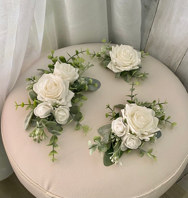 K&ouml;nnte beinhalten: Drei Blumenarrangements bestehend aus wei&szlig;en Rosen und kleineren Rosenknospen, akzentuiert mit gr&uuml;nen Eukalyptusbl&auml;ttern und Zweigen. Die Blumen sind in Clustern mit weichem, salbeifarbenem Laub angeordnet und bilden ein dekoratives Set.