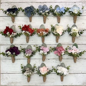 May include: Twenty assorted artificial flower buttonholes with greenery and twine accents. The buttonholes are arranged in four rows of five, with each row featuring a different colour scheme. The colours include blue, red, orange, white, purple, pink, and grey.