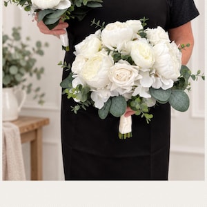 May include: Two artificial floral bouquets featuring white roses, peonies, and hydrangeas accented with green eucalyptus leaves. Each bouquet has a handle wrapped in white satin ribbon. The flowers are arranged in a rounded, full shape.
