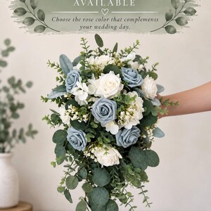 Op de afbeelding: Een watervalboeket met stoffige blauwe en witte rozen, afgewisseld met kleine witte vulbloemen en diverse groene eucalyptusbladeren. De tekst bovenaan luidt: CUSTOM ROSE COLORS AVAILABLE. Choose the rose color that complements your wedding day.