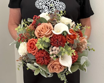 May include: A bridal bouquet featuring a variety of flowers in shades of orange, peach, and white. The bouquet includes roses, calla lilies, and other greenery. The text "TallulahRoseArtistry.com" is printed on the black shirt.