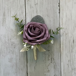 Op de afbeelding: Een corsage met een enkele stoffig paarse schuimroos als middelpunt, geaccentueerd met kleine takjes groene eucalyptusbladeren en piepkleine witte vulbloemen. De steel is omwikkeld met een cr&egrave;mekleurig lint met een kleine strik aan de basis.
