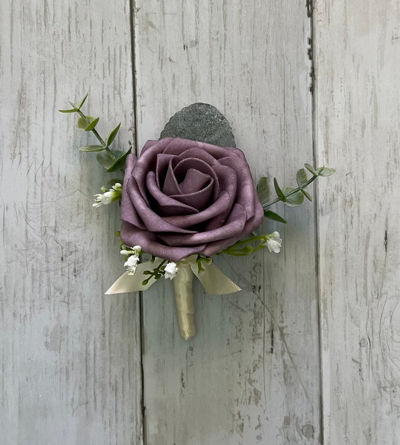 Op de afbeelding: Een corsage met een enkele stoffig paarse schuimroos als middelpunt, geaccentueerd met kleine takjes groene eucalyptusbladeren en piepkleine witte vulbloemen. De steel is omwikkeld met een cr&egrave;mekleurig lint met een kleine strik aan de basis.