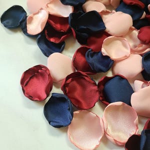 May include: A pile of silk rose petals in burgundy, navy blue, and blush pink colors. The petals are scattered on a white surface.