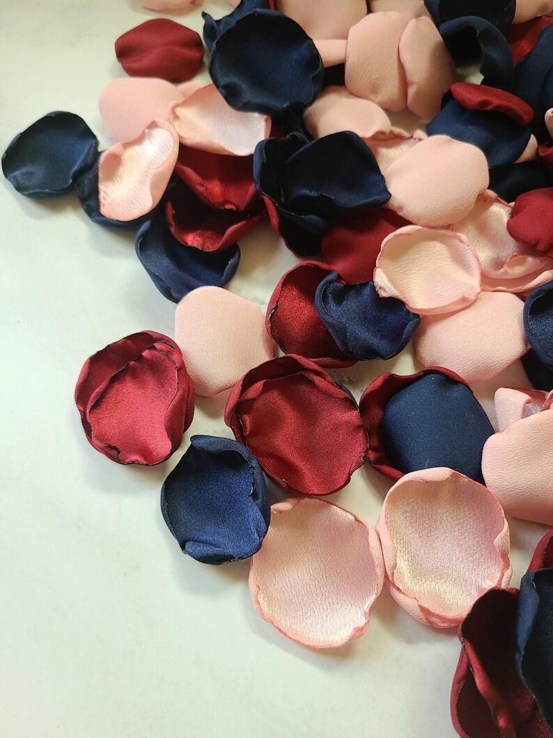 May include: A pile of silk rose petals in burgundy, navy blue, and blush pink colors. The petals are scattered on a white surface.