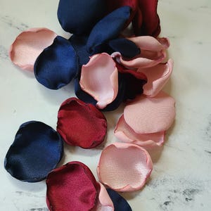 May include: Assortment of silk rose petals in navy blue, burgundy, and blush pink colors. The petals are scattered on a white marble surface.