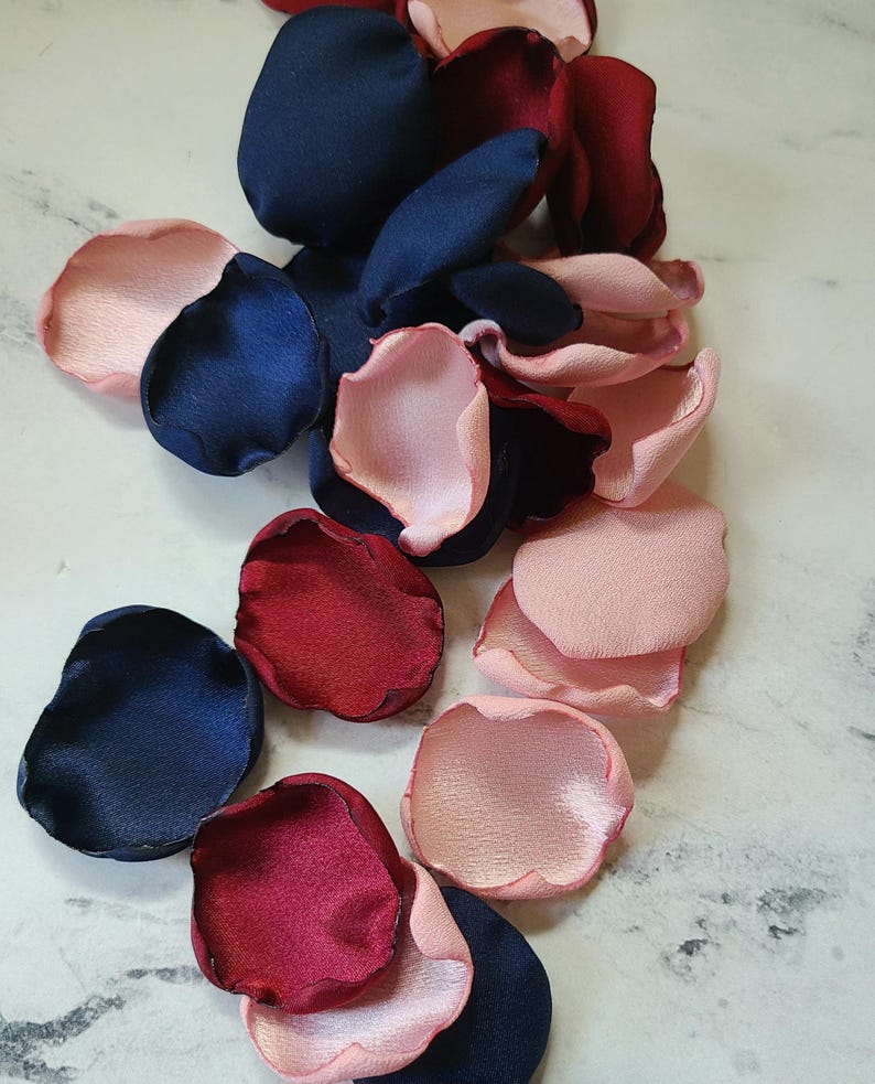 May include: Assortment of silk rose petals in navy blue, burgundy, and blush pink colors. The petals are scattered on a white marble surface.