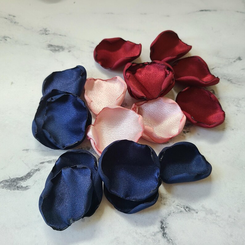 May include: A collection of silk rose petals in navy blue, blush pink, and burgundy. The petals are scattered on a white marble surface.