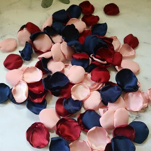 May include: A pile of silk rose petals in navy blue, burgundy, and blush pink colors. The petals are scattered on a white surface.