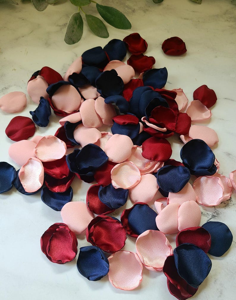 May include: A pile of silk rose petals in navy blue, burgundy, and blush pink colors. The petals are scattered on a white surface.