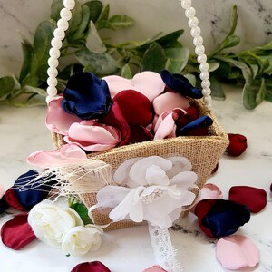 May include: A small burlap basket with a pearl handle filled with pink, burgundy, and navy blue silk flower petals. The basket is decorated with a white flower and a feather.