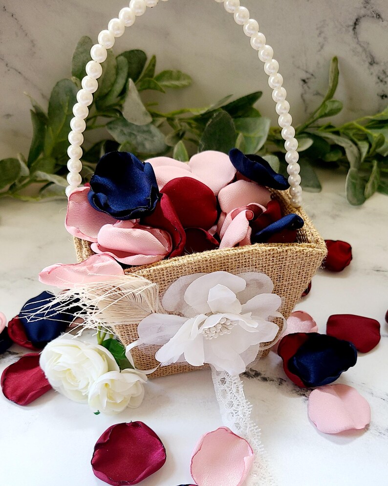 May include: A small burlap basket with a pearl handle filled with pink, burgundy, and navy blue silk flower petals. The basket is decorated with a white flower and a feather.
