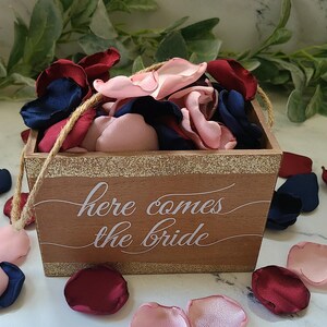 May include: A wooden box with a gold glitter trim and the text "here comes the bride" in white lettering. The box is filled with pink, burgundy, and navy blue fabric flower petals.